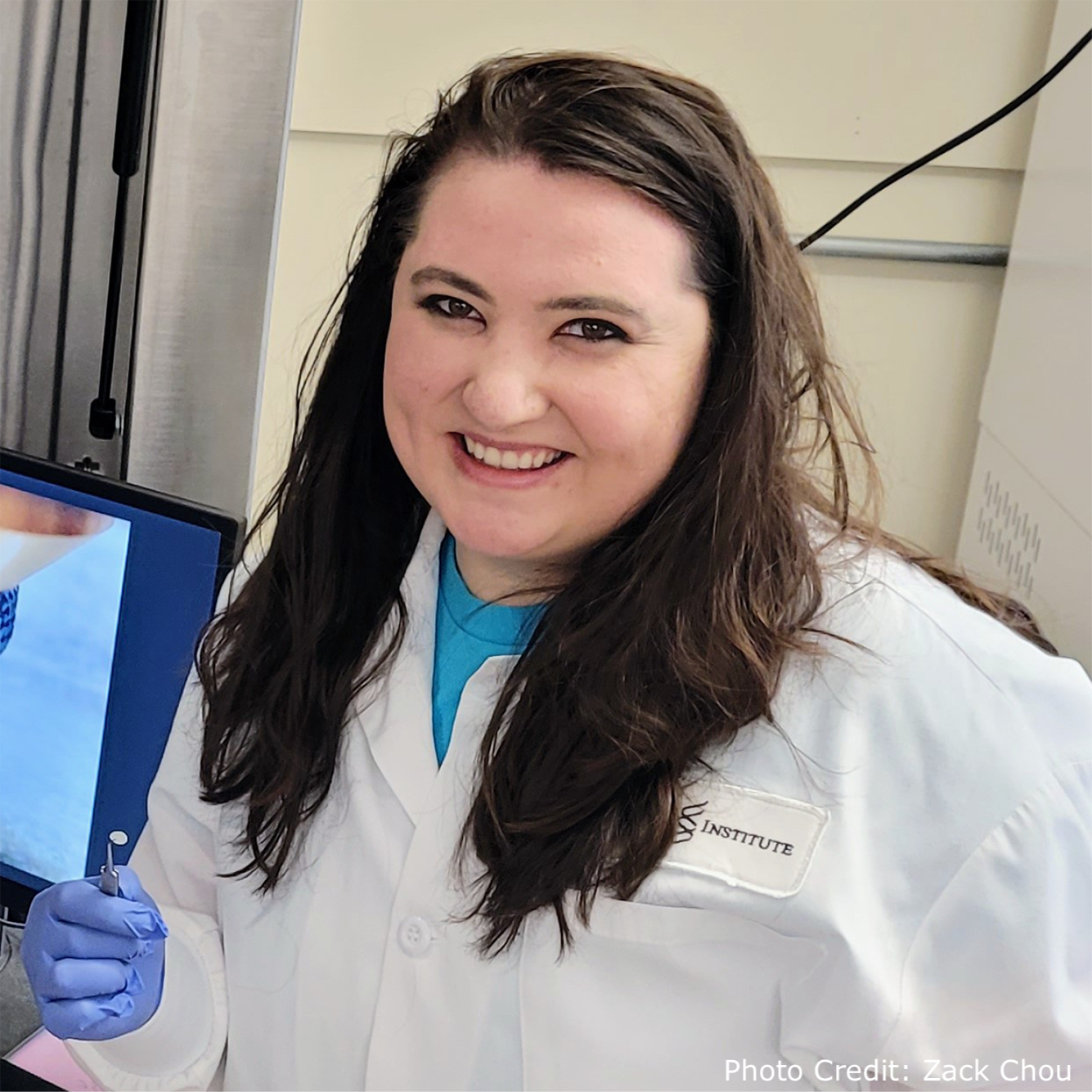 Harvard University graduate student Nicole Black in the lab, in front of a computer monitor that is displaying a picture of her invention being 3D printed. She also holds her small, biodegradable eardrum graft invention in tweezers.
