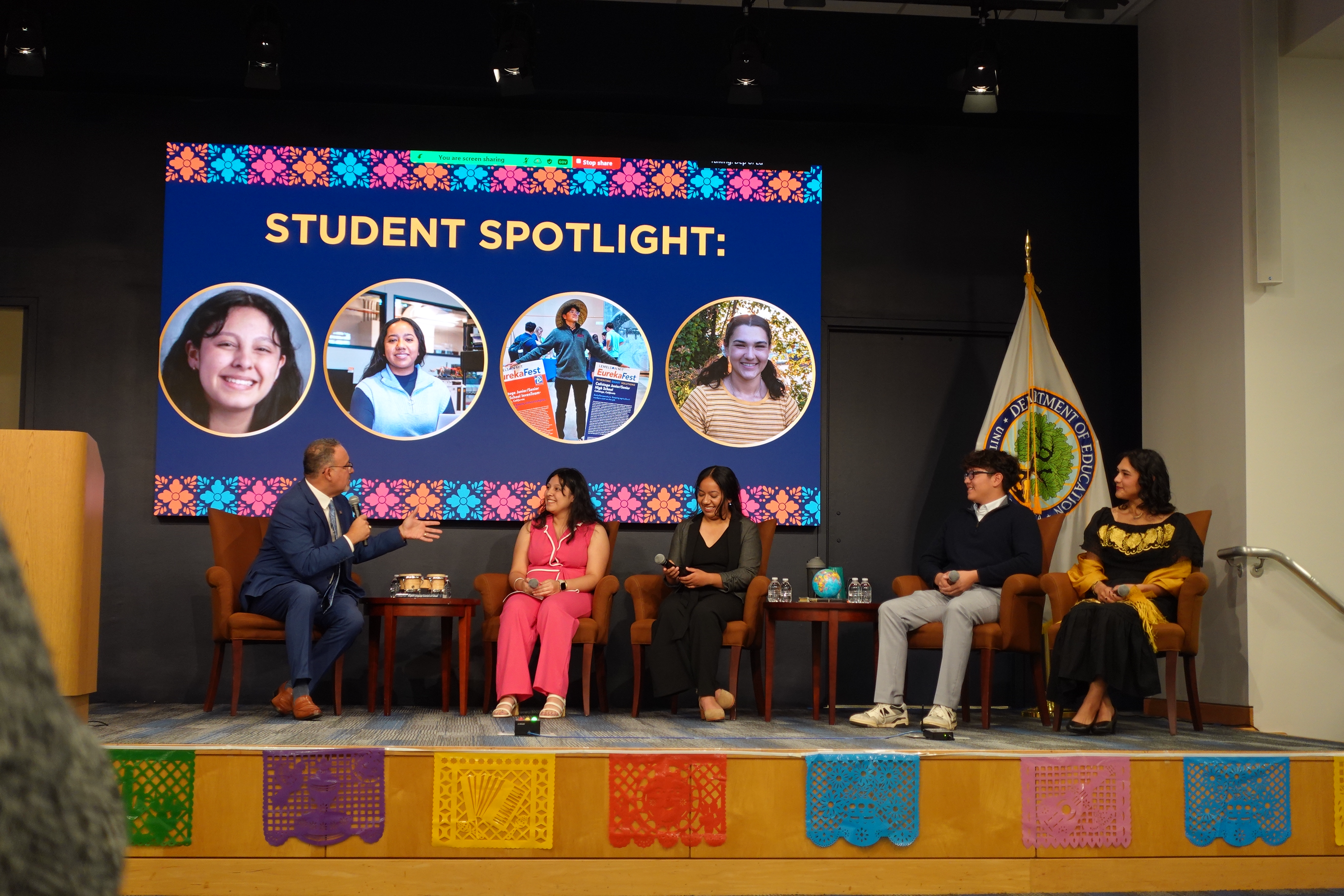 U.S. Secretary of Education Miguel Cardona is sitting on the right of the stage holding a microphone. He is leading a panel discussion with four student inventors seated nearby. Next to him is a young female inventor, then LMIT InvenTeam alum Lesly Rojas, and Calistoga InvenTeam member Elias Escobar, and InvenTeam alum Katia Avila. At the front of the stage floor, there are colorful flags, and behind the panelists is a big screen that reads “Student Spotlight” and shows pictures of the panelists. The U.S. Department of Education flag is also on display behind them.