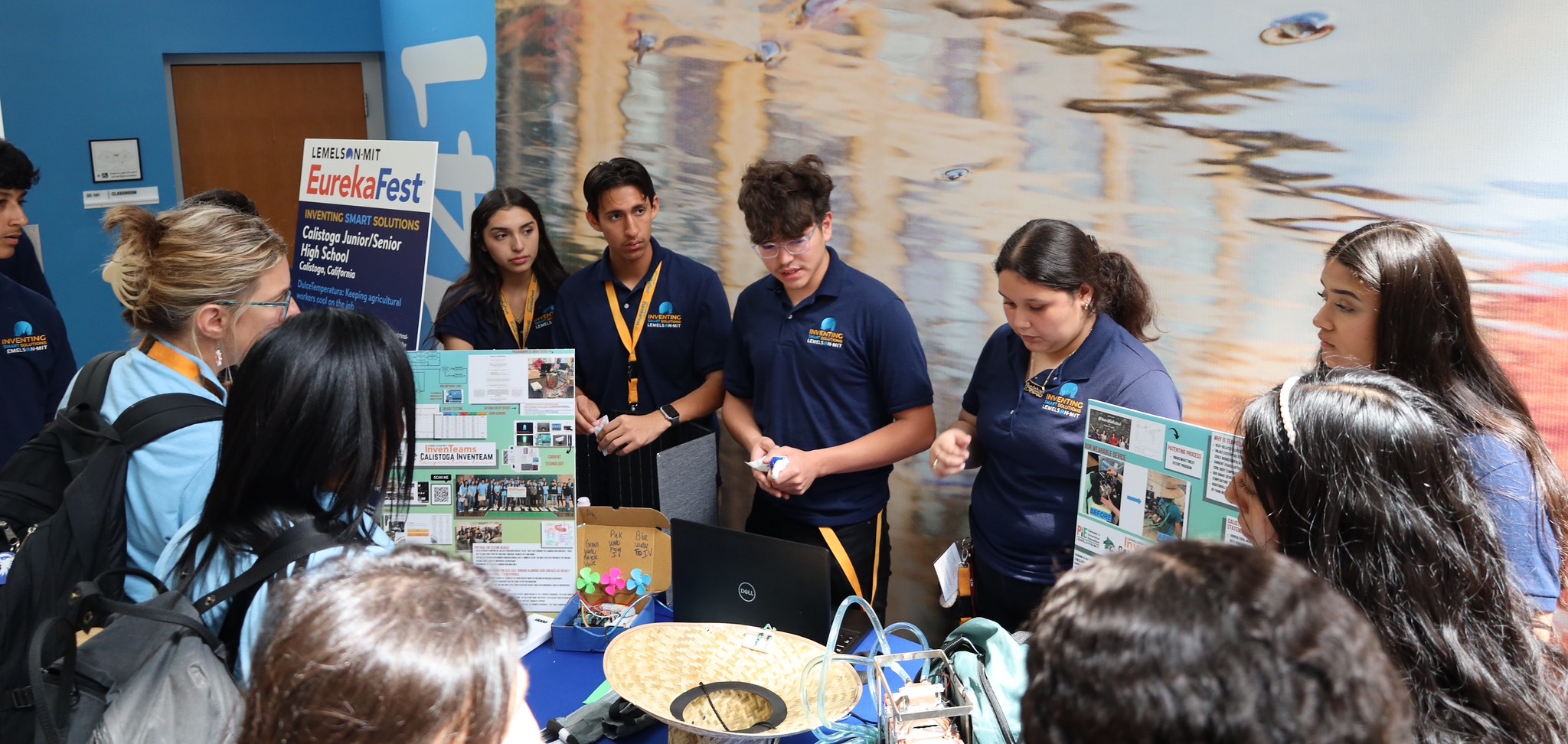 Five students from the Calistoga InvenTeam are standing behind a table, showing their invention project to other students at EurekaFest. There are posters and other materials related to their project on the table in front of them.