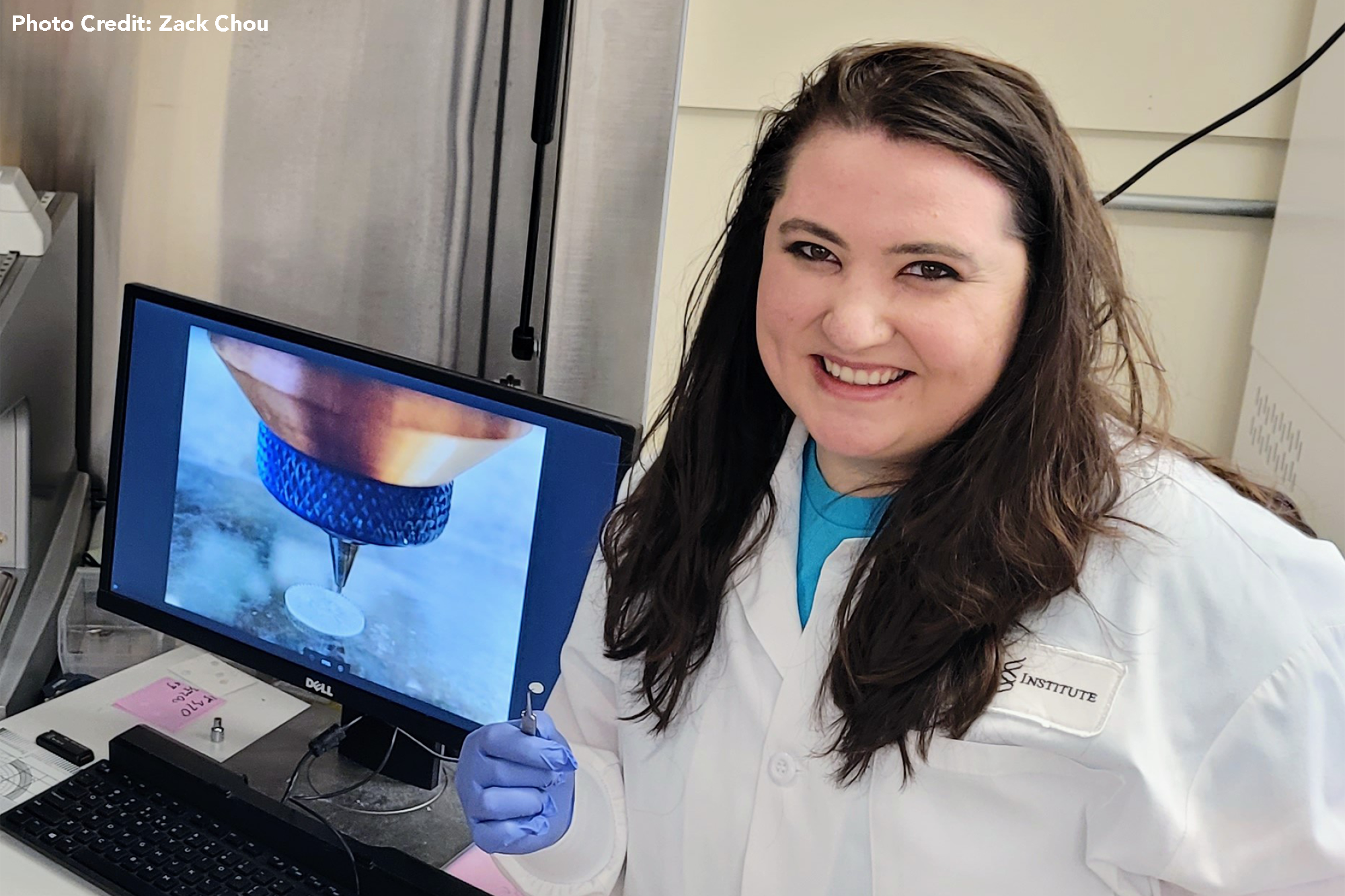 Harvard University graduate student Nicole Black in the lab, infront of a computer monitor that is displaying a picture of her inventionbeing 3D printed. She also holds her small, biodegradable eardrum graft invention in tweezers.
