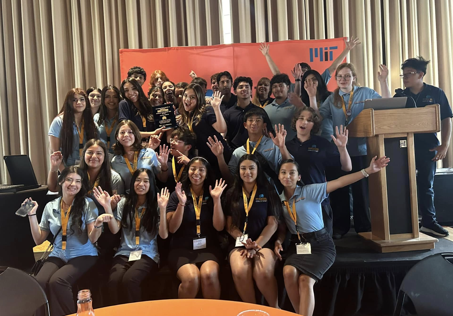 A group of smiling students wearing Lemelson-MIT branded shirts are standing and sitting together on stage with a MIT banner behind them. One of the students holds up a plaque award while others have their arms raised in celebration.