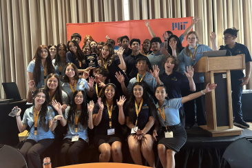 A group of smiling students wearing Lemelson-MIT branded shirts are standing and sitting together on stage with a MIT banner behind them. One of the students holds up a plaque award while others have their arms raised in celebration.
